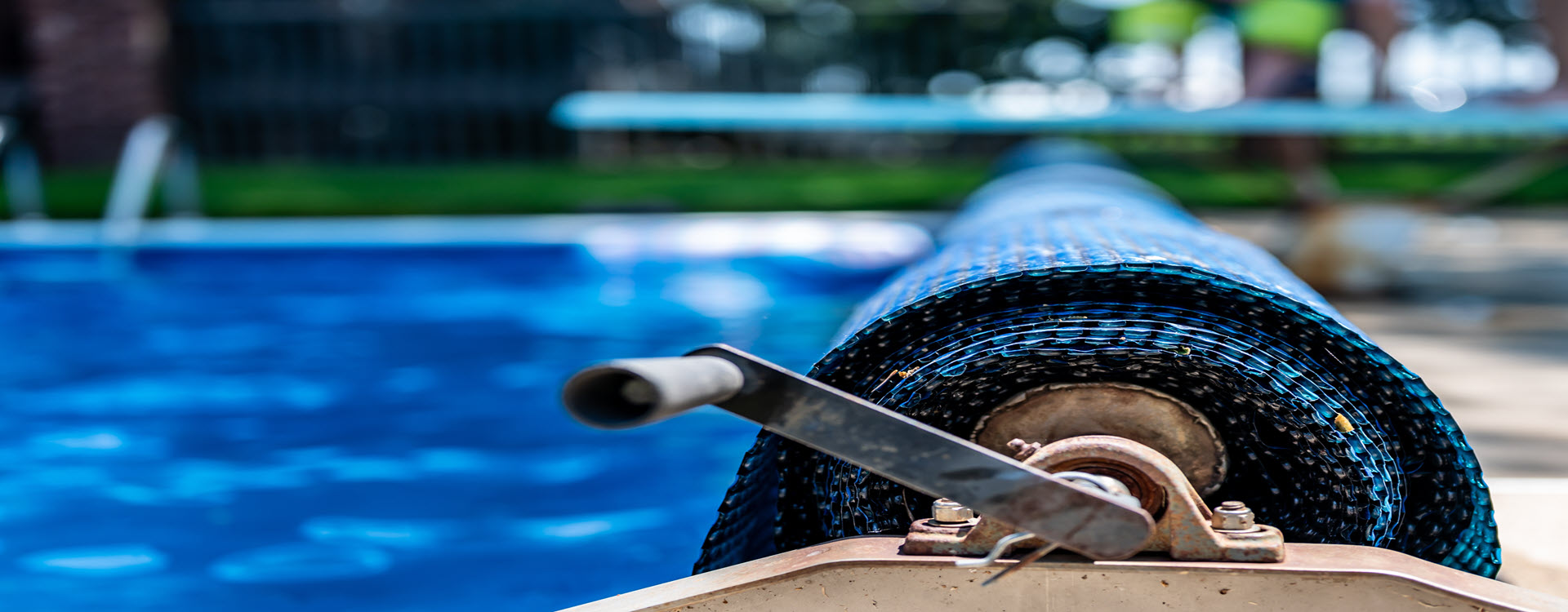 Close-up of a pool cover reel with a rolled-up blue solar cover beside a clear blue swimming pool.