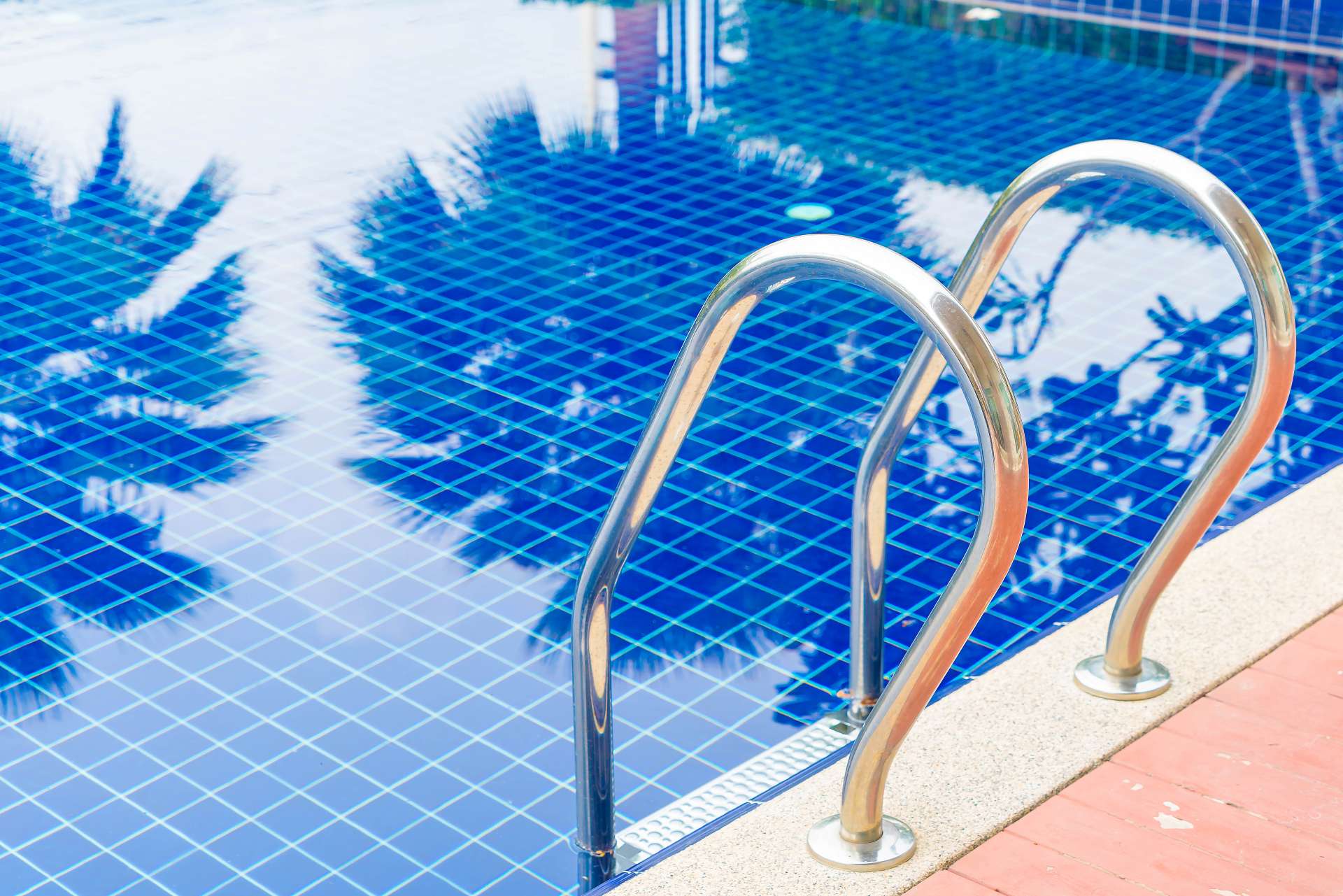 Close-up of a clean blue swimming pool with stainless steel handrails reflecting palm trees.
