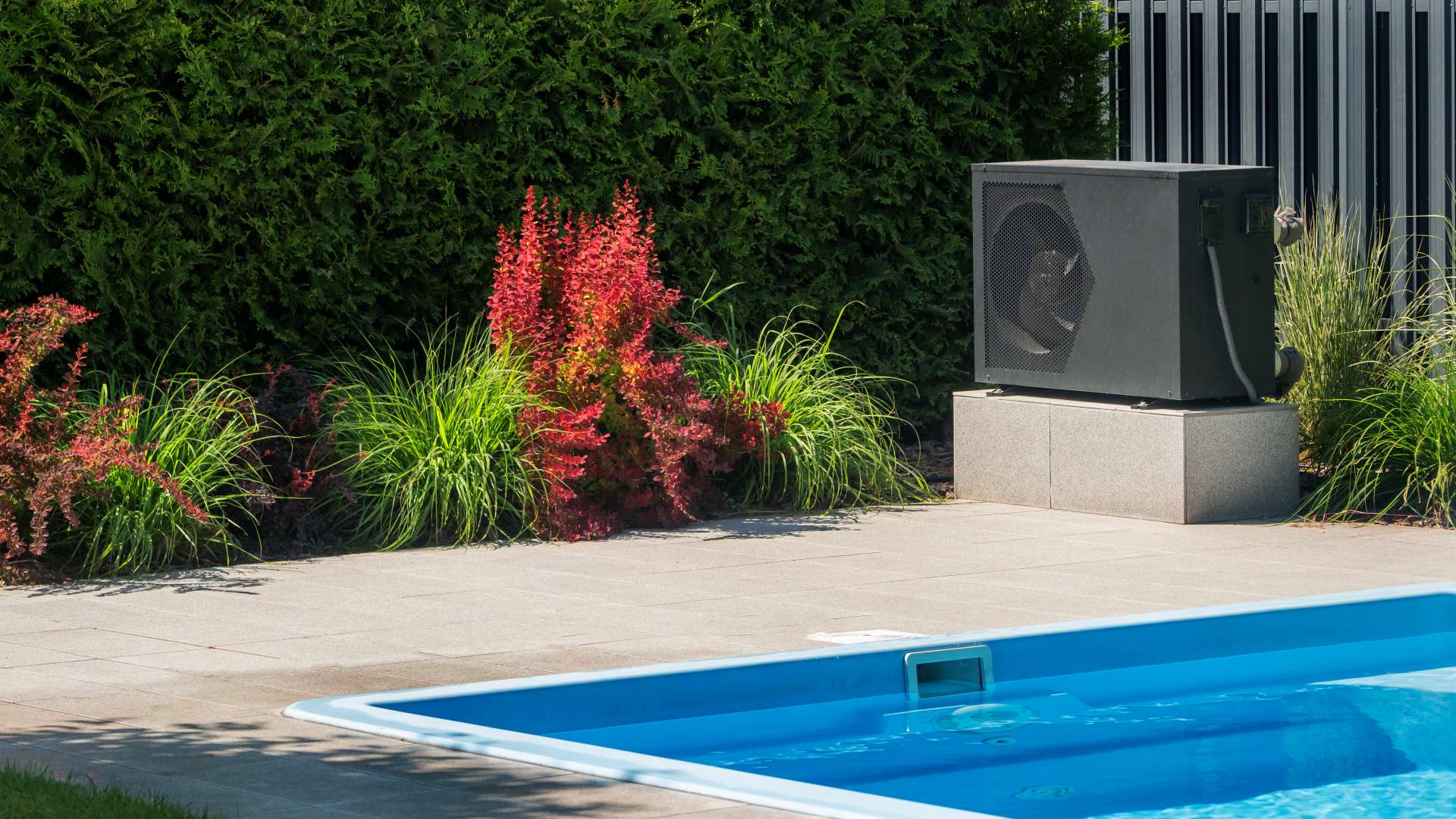 Modern pool heat pump unit installed near a blue swimming pool surrounded by decorative garden plants.