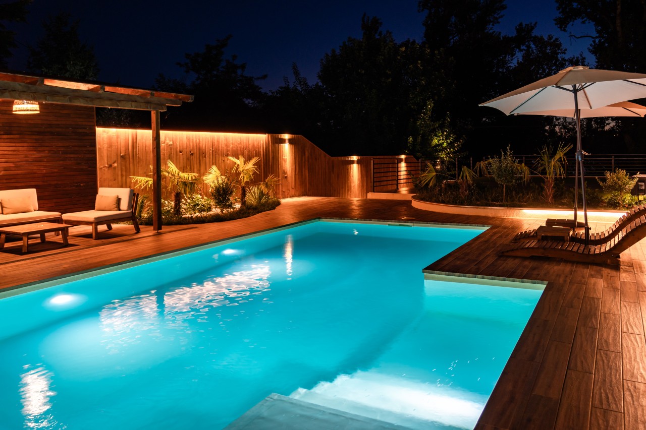 Swimming pool at night with bright underwater lighting and a wooden deck lounge space.