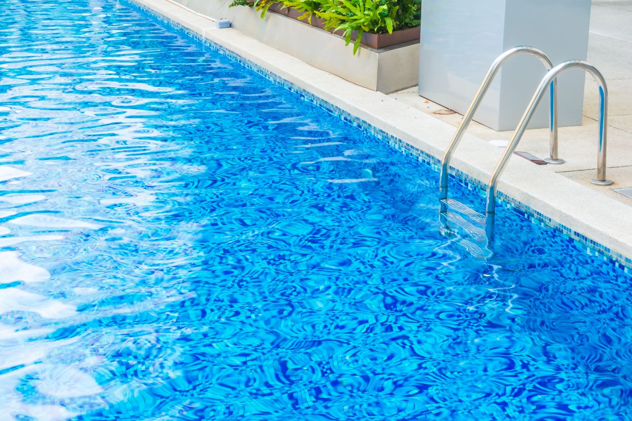 Bright blue swimming pool with clean, intact waterline tiles and stainless steel ladder beside a well-maintained pool deck.