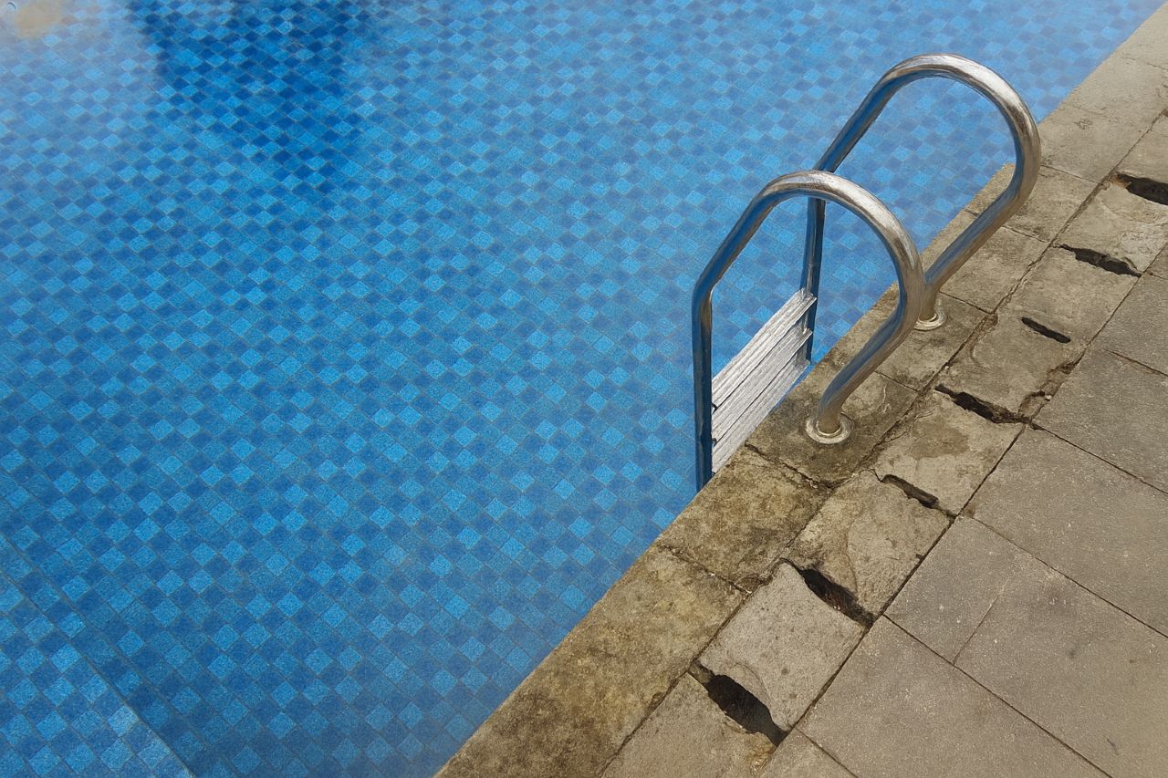 Old pool edge showing damaged coping stones and worn mosaic tile near the stainless steel ladder.