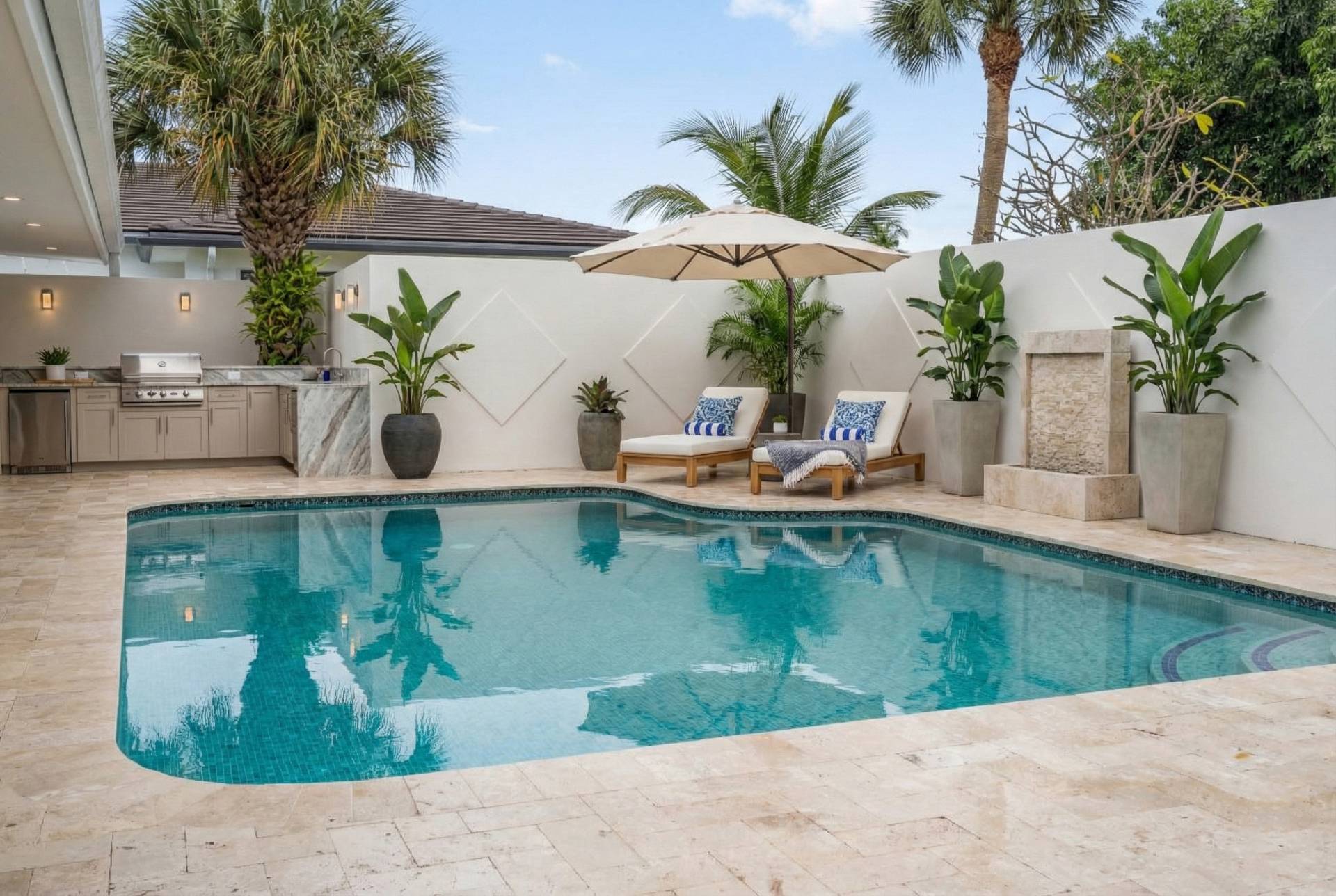 A beautiful backyard scene featuring a large swimming pool with turquoise water and travertine paver coping. Two lounge chairs with blue and white cushions are placed under a beige umbrella next to the pool. A fully equipped outdoor kitchen with a grill, sink, and light cabinetry is visible in the background against a white wall, surrounded by lush tropical plants and palm trees under a clear blue sky.